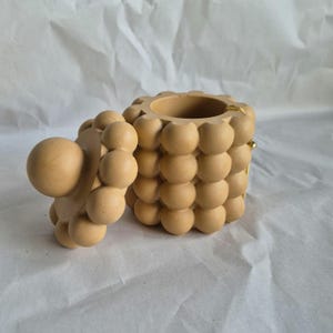 May include: A beige, spherical-shaped jewelry box with a lid. The box and lid are covered in a bubble-like pattern. The lid is open, revealing the interior. The box is on a white surface.