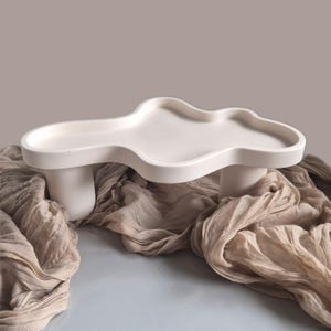 May include: A white, cloud-shaped decorative tray with a raised edge, supported by two cylindrical legs. The tray is made of a smooth material and sits on a beige fabric. The overall design is minimalist and modern.
