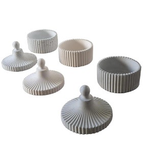 Peut inclure: Ensemble de cinq contenants en béton blanc avec couvercles. Les contenants ont un design côtelé et les couvercles ont une forme de dôme avec un bouton sur le dessus.