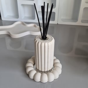 May include: A white, ribbed ceramic diffuser with black reeds. The diffuser sits on a round, white, bubble-shaped base. The design is minimalist and modern, suitable for home fragrance.