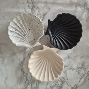May include: Three decorative seashell-shaped dishes. One is white with black speckles, one is black, and one is a light beige.