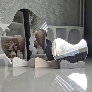 Peut inclure: Trois miroirs décoratifs aux formes uniques, chacun monté sur une base texturée. Les miroirs reflètent l'environnement, avec une palette de couleurs neutres et une esthétique moderne. Les bases sont dans des tons de gris et de beige.