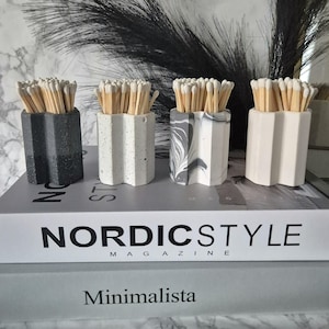 May include: Four decorative matchstick holders made of concrete in black, white with black speckles, white with grey marbling, and white. Each holder has a scalloped design and is filled with wooden matchsticks with white tips. The holders are sitting on a stack of magazines with the text "Nordics Style Magazine" and "Minimalista" visible.