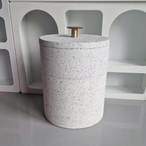 May include: A cylindrical, speckled white container with a matching lid. The lid is topped with a small, gold-colored, T-shaped handle. The container is likely for storage and is made of a textured material.