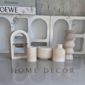 Könnte beinhalten: Eine Sammlung von Wohnaccessoires in neutralen Farbtönen. Die Anordnung umfasst ein kleines bogenförmiges Objekt mit einer Metallschale, einen zylindrischen Behälter, eine Schachtel mit Deckel, zwei Vasen mit strukturierten Oberflächen und eine hohe, wellenförmige Vase. Der Text "HOME DECOR" steht unten.