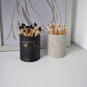 May include: Two cylindrical match holders, one black with gold marbling and the other white with grey marbling, each filled with matches. The matches in the black holder have black tips, while the matches in the white holder have natural tips.