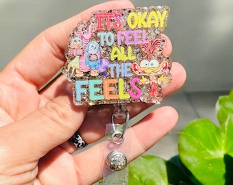 Könnte beinhalten: Ein goldener Glitzer-Badgehalter mit einem bunten Cartoon-Bild und dem Text "It's okay to feel all the feels".