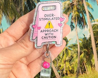 May include: A pink glittery badge reel shaped like a caution sign with the text OVER-STIMULATED APPROACH WITH CAUTION and a yellow warning triangle. It features small pink hearts on the sides and a clear strap with a small pink bead and heart charm.