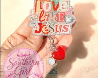 May include: A clear badge reel with the words "Love Like Jesus" in red, pink, and white. The badge has a red heart charm and a silver retractable cord. The badge is held in a hand.