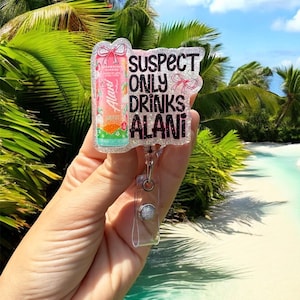 Pu&ograve; includere: Un portabadge trasparente con dettagli glitterati e la scritta "SUSPECT ONLY DRINKS ALANI". Il badge presenta un design di bevanda Alani Nu rosa e arancione. Il portabadge &egrave; tenuto in mano su uno sfondo tropicale.