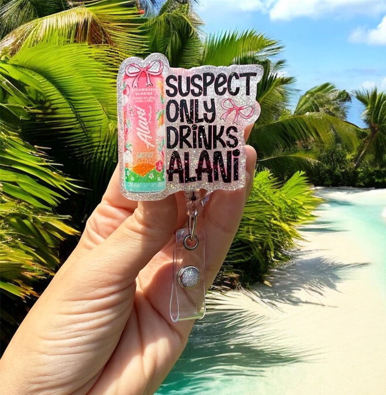 Pu&ograve; includere: Un portabadge trasparente con dettagli glitterati e la scritta "SUSPECT ONLY DRINKS ALANI". Il badge presenta un design di bevanda Alani Nu rosa e arancione. Il portabadge &egrave; tenuto in mano su uno sfondo tropicale.