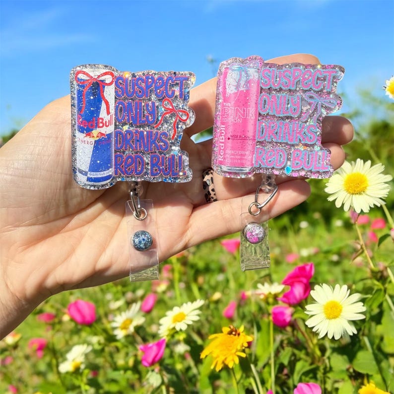 Pu&ograve; includere: Due porta badge glitterati con disegni di lattine Red Bull. Uno presenta una lattina blu, l'altro una lattina rosa. Il testo recita "SUSPECT ONLY DRINKS RED BULL" in glitter viola e argento. Ogni porta badge ha un cinturino in plastica trasparente e un bottone decorativo.
