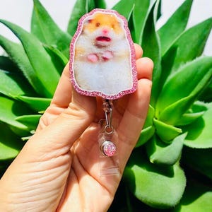 May include: A pink glitter-edged badge reel featuring a cartoon hamster design. The hamster has a white body, orange and brown fur, and pink facial features. The badge reel has a clear plastic strap and a pink glitter button.