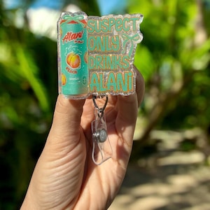 Pu&ograve; includere: Un portabadge glitterato. Il portabadge presenta una lattina turchese di Alani Nu Peach, con la scritta "SUSPECT ONLY DRINKS ALANI" in lettere turchesi. Il portabadge ha una clip in plastica trasparente.