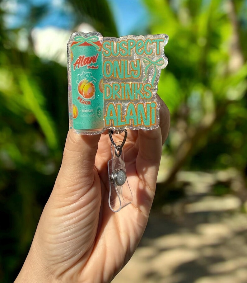 Pu&ograve; includere: Un portabadge glitterato. Il portabadge presenta una lattina turchese di Alani Nu Peach, con la scritta "SUSPECT ONLY DRINKS ALANI" in lettere turchesi. Il portabadge ha una clip in plastica trasparente.