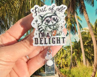 May include: A glittery acrylic badge reel featuring a black and white illustration of a raccoon wearing heart-shaped sunglasses and holding a drink. The text reads First of all I'M A DELIGHT. The badge reel has a clear plastic strap and a heart charm.