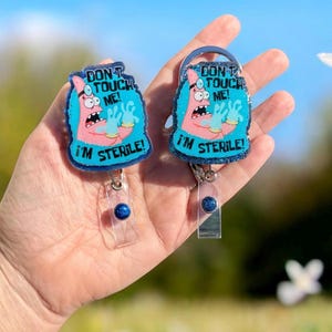 May include: Two badge reels featuring a pink cartoon character wearing a head mirror and blue gloves on a glittery blue background. The text reads "DON'T TOUCH ME! I'M STERILE!" in black lettering. Each reel has a clear plastic strap and a blue button.