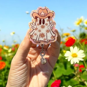 May include: A badge reel featuring a cartoon ghost wearing a cowboy hat with a star and peace signs. The ghost is decorated with floral patterns and outlined in brown. The badge reel has a clear plastic strap and a silver-colored charm.