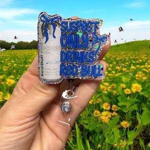 Pu&ograve; includere: Un portabadge glitterato e trasparente con un design a lattina di Red Bull e la scritta "Suspect Only Drinks Red Bull" in blu. Il portabadge ha un supporto in plastica trasparente e una clip color argento.