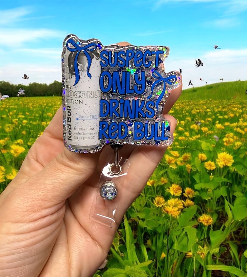 Pu&ograve; includere: Un portabadge glitterato e trasparente con un design a lattina di Red Bull e la scritta "Suspect Only Drinks Red Bull" in blu. Il portabadge ha un supporto in plastica trasparente e una clip color argento.