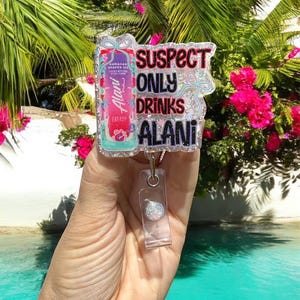 Pu&ograve; includere: Un portabadge con la scritta "SUSPECT ONLY DRINKS ALANI" in glitter neri, rosa e verde acqua. Il portabadge presenta anche un'immagine colorata di una lattina di bevanda energetica Alani Nu. Lo sfondo &egrave; una scena tropicale.