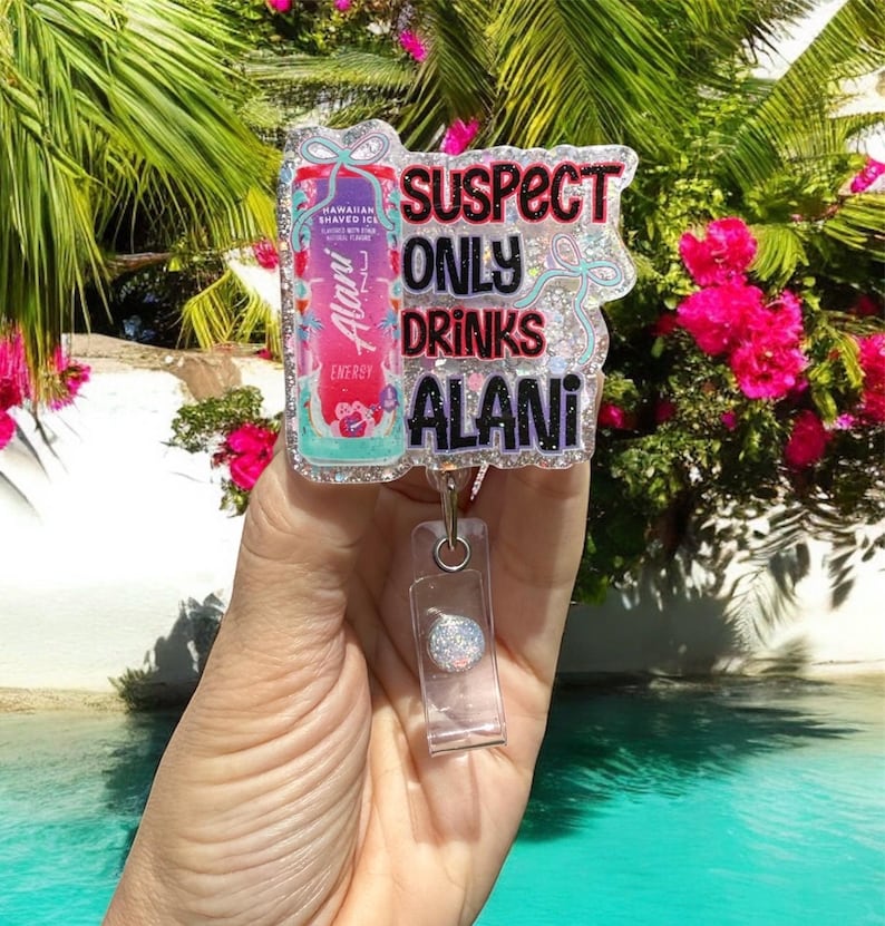 Pu&ograve; includere: Un portabadge con la scritta "SUSPECT ONLY DRINKS ALANI" in glitter neri, rosa e verde acqua. Il portabadge presenta anche un'immagine colorata di una lattina di bevanda energetica Alani Nu. Lo sfondo &egrave; una scena tropicale.