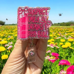 Pu&ograve; includere: Un portabadge rosa glitterato con la scritta "SUSPECT ONLY DRINKS RED BULL" e un design a lattina Red Bull. Il portabadge ha un supporto in plastica trasparente e un cordino retrattile. Lo sfondo &egrave; un campo di fiori gialli e rosa.