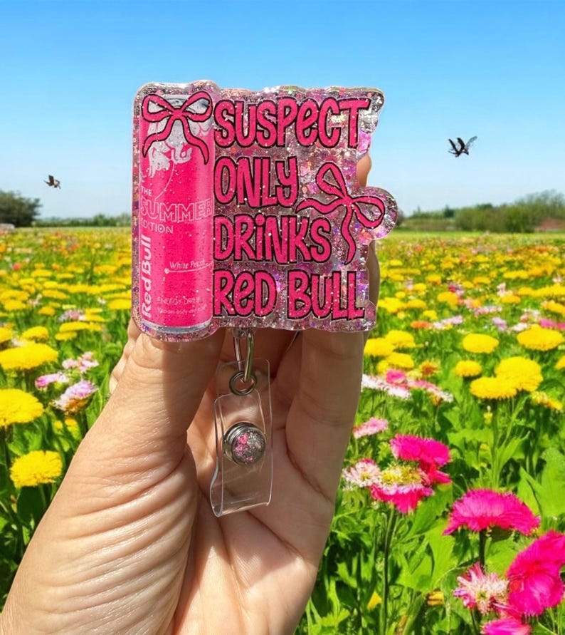 Pu&ograve; includere: Un portabadge rosa glitterato con la scritta "SUSPECT ONLY DRINKS RED BULL" e un design a lattina Red Bull. Il portabadge ha un supporto in plastica trasparente e un cordino retrattile. Lo sfondo &egrave; un campo di fiori gialli e rosa.