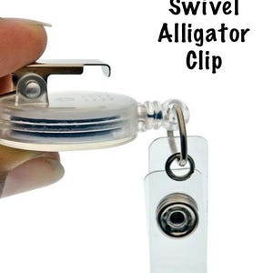 Pu&ograve; includere: Un portabadge con clip a coccodrillo trasparente e non girevole. Il portabadge ha una clip in metallo e una cinghia in plastica trasparente con chiusura a scatto. Il testo "Non Swivel Alligator Clip" &egrave; stampato in nero.