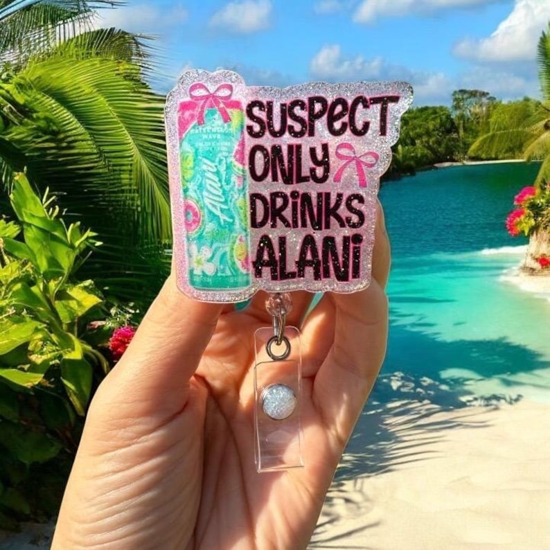 Pu&ograve; includere: Un portabadge glitterato con la scritta "SUSPECT ONLY DRINKS ALANI" in nero e rosa. Il badge presenta un design colorato di una lattina di bevanda. Il portabadge &egrave; tenuto da un supporto in plastica trasparente con un bottone argentato.