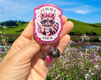 May include: A pink and glittery badge reel featuring a raccoon with heart-shaped sunglasses and a bow. The text on the badge reel reads "CUTE BUT FERAL". The reel has a black heart charm and a clear plastic ID holder.