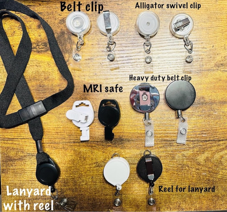 Pu&ograve; includere: Assortimento di porta badge e cordini. L'immagine mostra vari stili, tra cui clip da cintura, clip girevole a coccodrillo e modelli di clip da cintura per impieghi gravosi. Il testo "MRI safe" &egrave; visibile. &Egrave; presente anche un cordino nero con un avvolgitore.