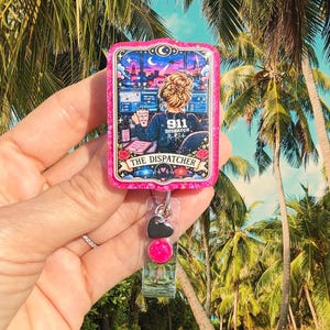 May include: A rectangular badge reel with a bright pink glitter border featuring a tarot card illustration titled THE DISPATCHER. The graphic shows a person with a messy bun at a computer desk with monitors, a coffee mug, and 911 DISPATCH text.