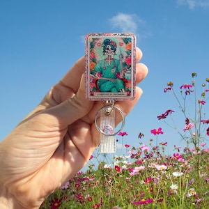 May include: A rectangular badge reel featuring a skeleton in a nurse's uniform, surrounded by flowers. The badge reel has a glittery border and a clear plastic strap with a metal ring. The text "LA MUERTE DE ENFERMERA" is at the bottom.