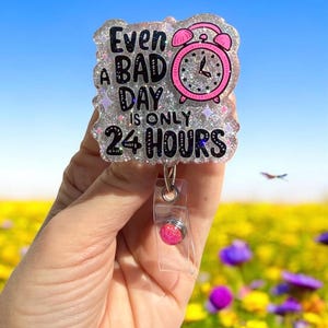 May include: A glittery badge reel with the text "Even a bad day is only 24 hours." The design includes a pink alarm clock and stars. The reel has a clear plastic strap and a pink gem-like button.