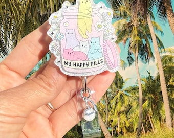 May include: A glittery acrylic badge reel featuring a jar illustration filled with five colorful cartoon cats. The jar has a pink lid and the text MY HAPPY PILLS printed on the front. The design is accented with small white flowers.