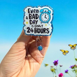 May include: A blue and white badge reel with the text "Even a bad day is only 24 hours" and a blue alarm clock graphic. The badge reel has a glittery border and a clear plastic clip with a blue bead.