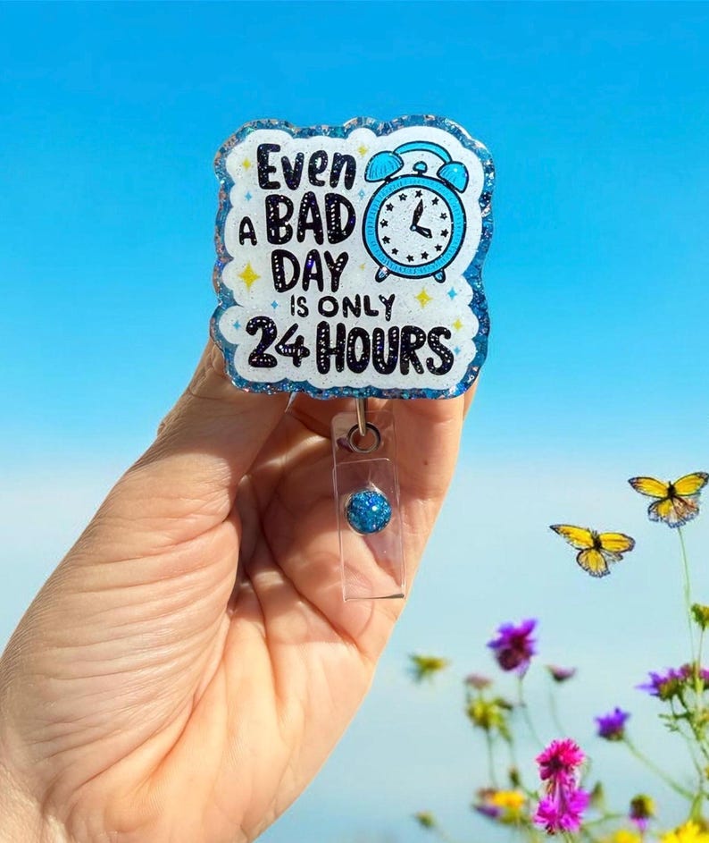 May include: A blue and white badge reel with the text "Even a bad day is only 24 hours" and a blue alarm clock graphic. The badge reel has a glittery border and a clear plastic clip with a blue bead.