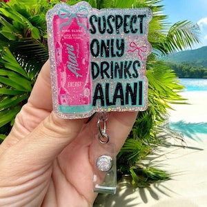 Pu&ograve; includere: Un portabadge glitterato con la scritta "SUSPECT ONLY DRINKS ALANI". Il badge presenta un design di bevanda energetica Alani Nu rosa e un avvolgitore in plastica trasparente con un bottone argentato. Lo sfondo mostra una scena di spiaggia tropicale.