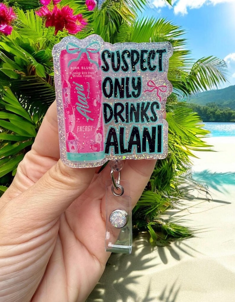 Pu&ograve; includere: Un portabadge glitterato con la scritta "SUSPECT ONLY DRINKS ALANI". Il badge presenta un design di bevanda energetica Alani Nu rosa e un avvolgitore in plastica trasparente con un bottone argentato. Lo sfondo mostra una scena di spiaggia tropicale.