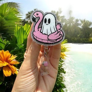 May include: A glittery badge reel featuring a cartoon ghost floating on a pink flamingo pool float. The badge reel has a clear plastic clip and a pink and silver accent. The background shows a tropical beach scene.