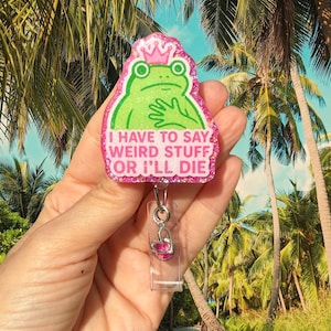 May include: A glittery pink badge reel featuring a green frog wearing a pink crown. The text reads, I HAVE TO SAY WEIRD STUFF OR I'LL DIE. A small silver heart charm hangs from the metal clip, which is attached to a clear plastic strap.