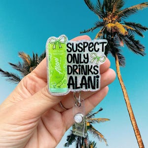 Pu&ograve; includere: Un porta badge trasparente con un design glitterato. Il badge presenta una lattina verde di bevanda energetica Alani Nu e il testo "SUSPECT ONLY DRINKS ALANI" in nero. Fiocchi verdi accentuano il design. Lo sfondo mostra un cielo blu e palme.