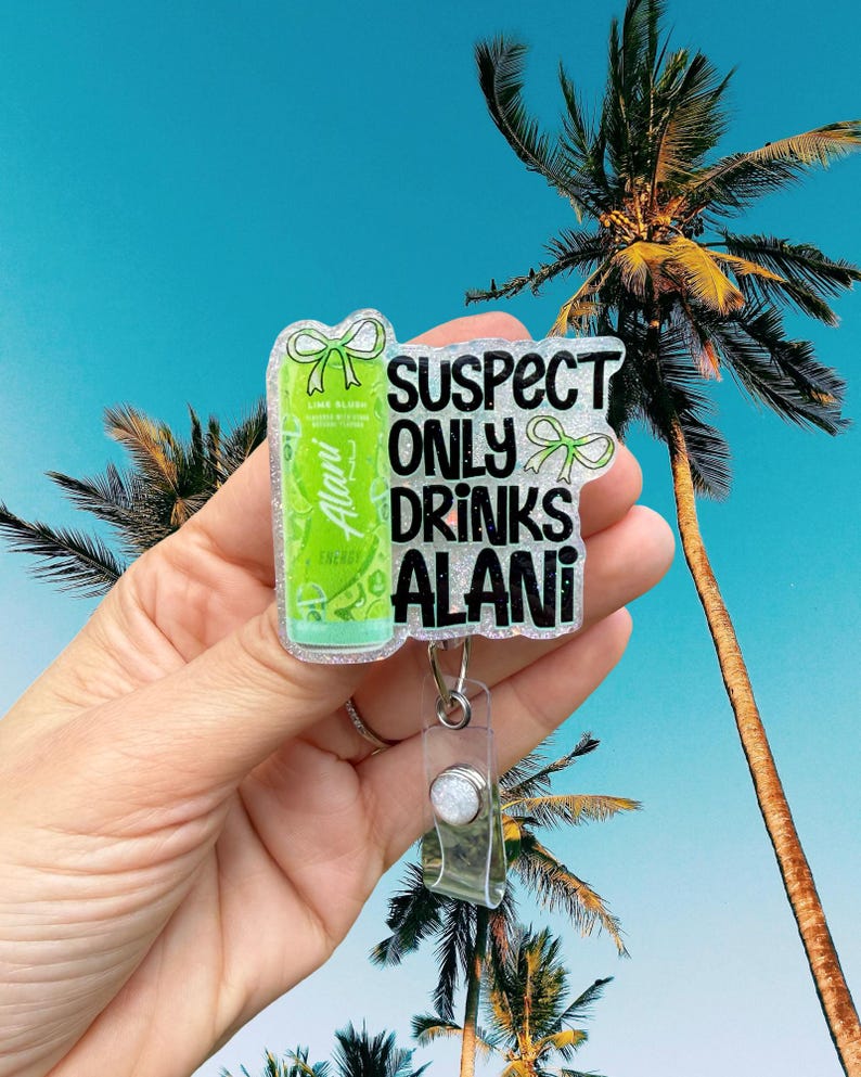 Pu&ograve; includere: Un porta badge trasparente con un design glitterato. Il badge presenta una lattina verde di bevanda energetica Alani Nu e il testo "SUSPECT ONLY DRINKS ALANI" in nero. Fiocchi verdi accentuano il design. Lo sfondo mostra un cielo blu e palme.