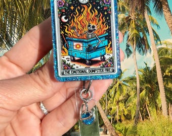 May include: A rectangular badge reel featuring a blue glitter frame and a tarot-style card illustration of a skeleton in a burning dumpster. The card includes the text ST and THE EMOTIONAL DUMPSTER FIRE. A silver heart charm hangs from the clear strap.