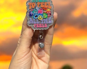 May include: A silver glitter badge holder with a clear retractable badge reel. The badge holder features a colorful image of the characters from the movie Inside Out with the text "It's okay to feel all the feels".