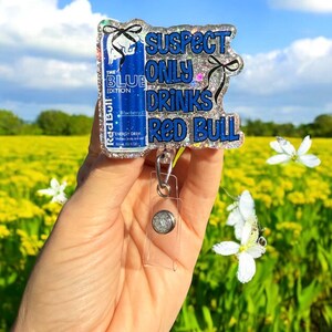 Pu&ograve; includere: Un portabadge glitterato con la scritta "SUSPECT ONLY DRINKS RED BULL". Il badge presenta un design di lattina Red Bull blu con le parole "THE BLUE EDITION". Il portabadge ha un supporto in plastica trasparente e un bottone argentato.