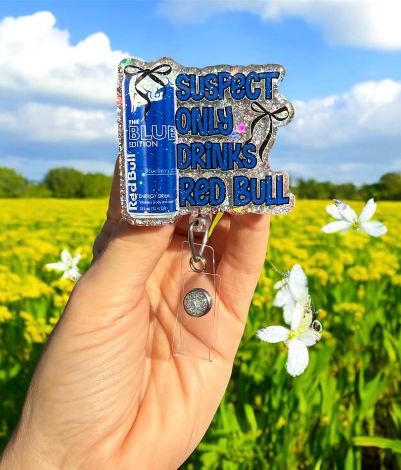 Pu&ograve; includere: Un portabadge glitterato con la scritta "SUSPECT ONLY DRINKS RED BULL". Il badge presenta un design di lattina Red Bull blu con le parole "THE BLUE EDITION". Il portabadge ha un supporto in plastica trasparente e un bottone argentato.