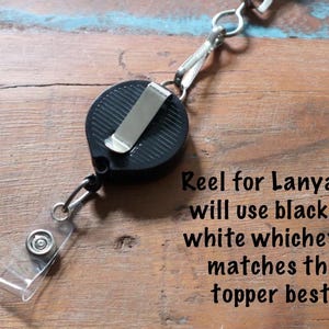 May include: Black retractable reel for a lanyard with a metal clip and clear plastic strap. The text on the image reads: "Reel for Lanyard: will use black or white whichever matches the topper best."