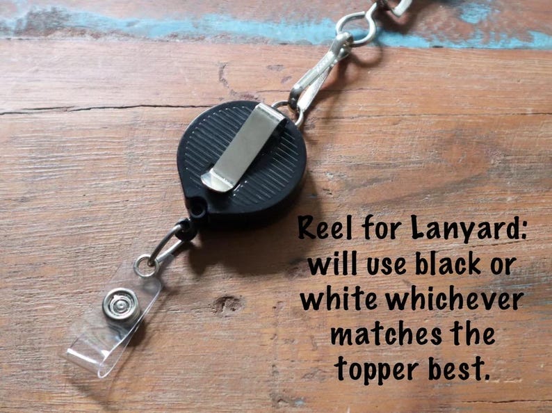 May include: Black retractable reel for a lanyard with a metal clip and clear plastic strap. The text on the image reads: "Reel for Lanyard: will use black or white whichever matches the topper best."